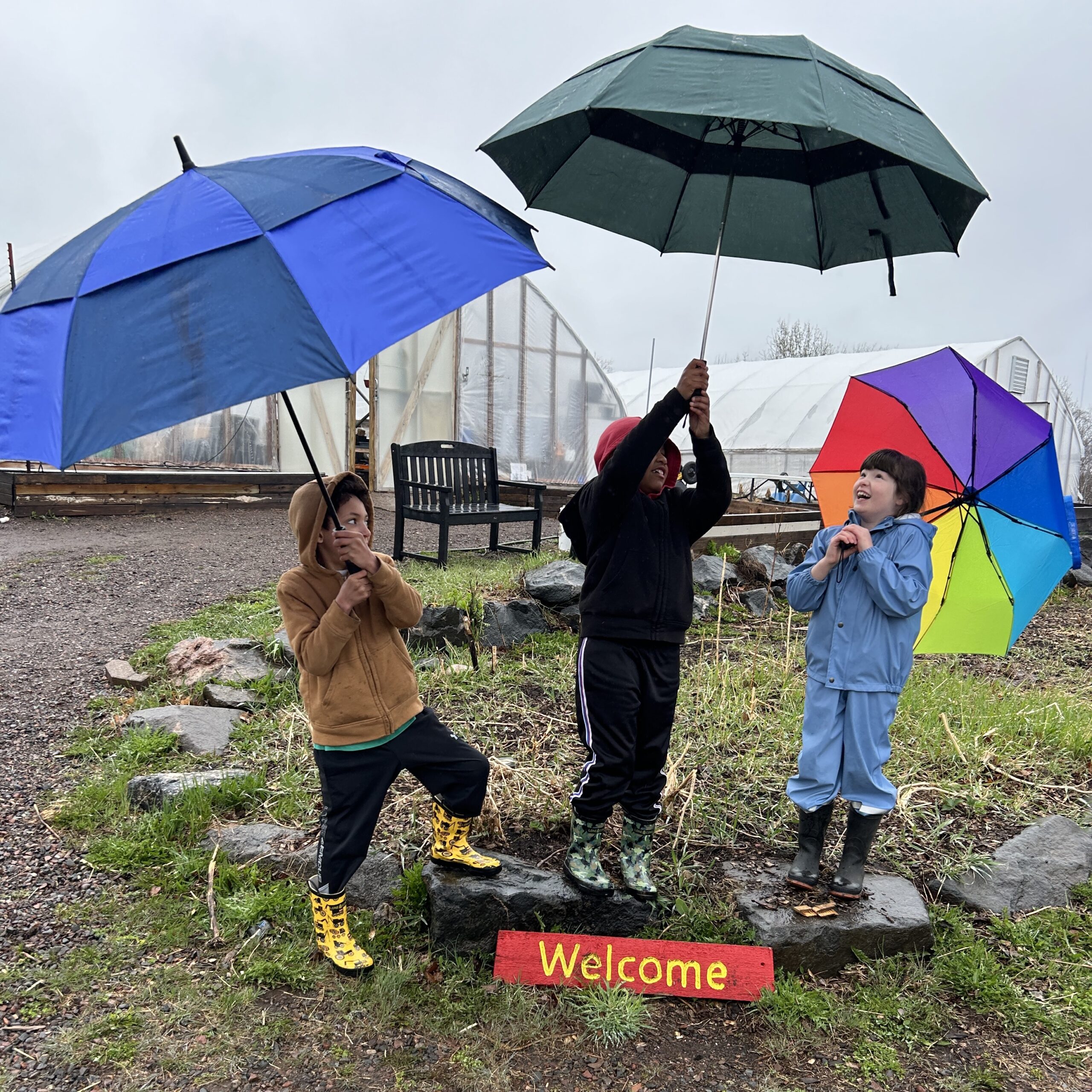 Three children with umbrellas pose joyfully by a "welcome" sign on the Farm.