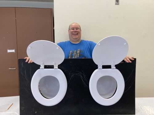 Patrick with toilet game 2023 Patrick smiles wide posing with finished fundraising game- two toilet seats attached to plywood.