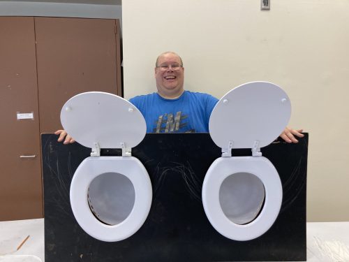 Patrick smiles wide posing with finished fundraising game- two toilet seats attached to plywood.