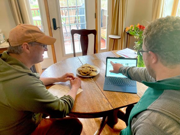 Two people sitting at a table together look at a computer screen. There are fresh-baked cookies on the table.